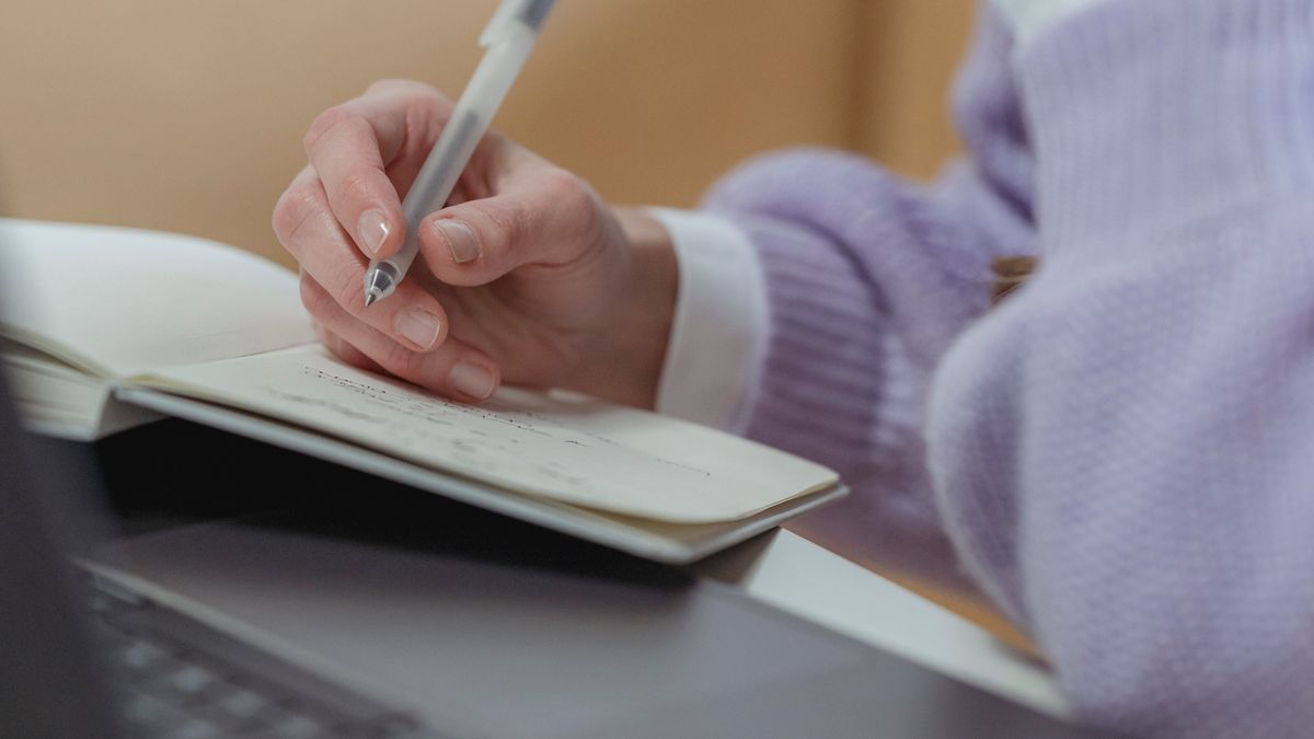 Person writing notes in a notebook next to a laptop